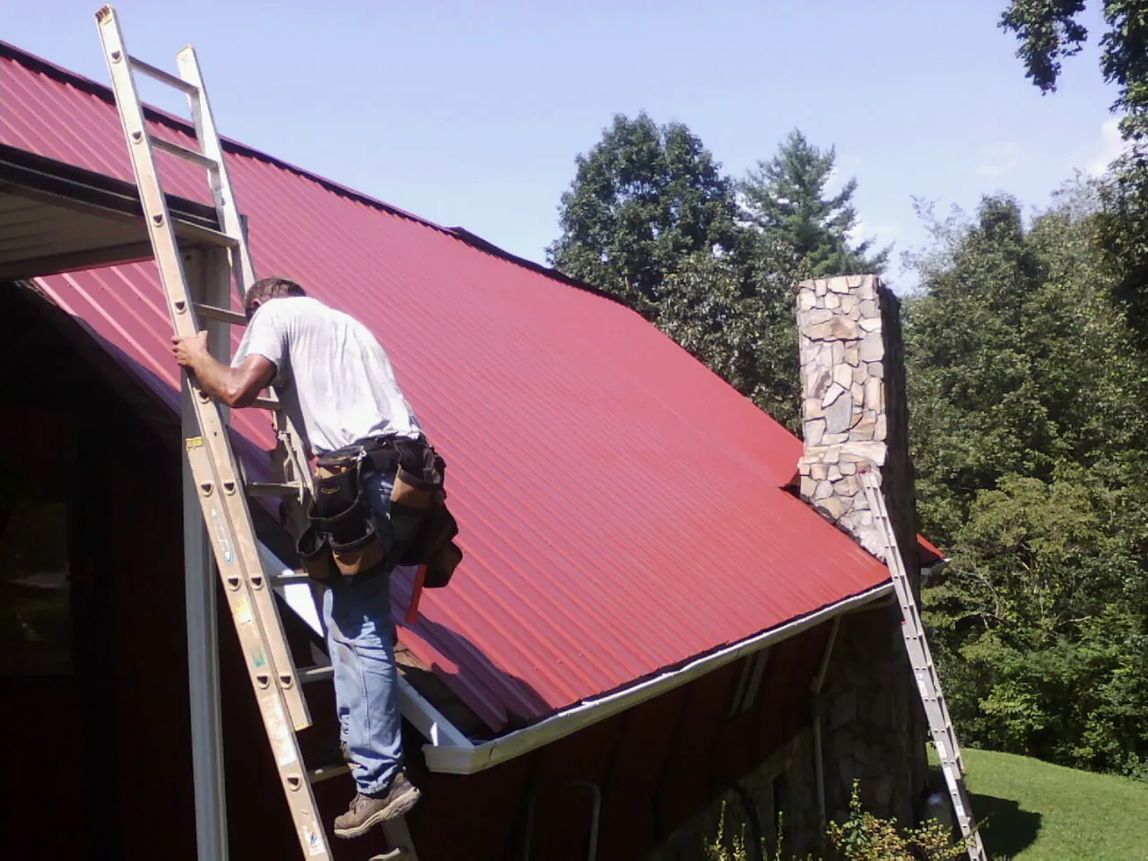 Roofer on a ladder beside a red metal roof and stone chimney — Soffit & Fascia Repair by Roofing Brothers in Doon
